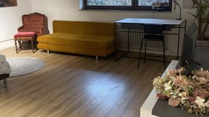 Una habitación con piso de madera, un sofá amarillo, un sillón rojo, un escritorio blanco y una ventana que da a zonas verdes.