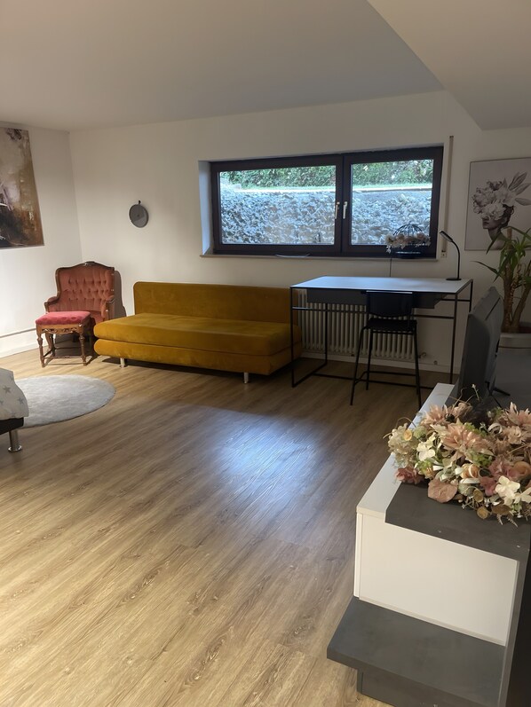 Une pièce avec un plancher en bois, un canapé jaune, un fauteuil rouge, un bureau blanc et une fenêtre donnant sur un paysage verdoyant.