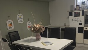 Un comedor con una mesa blanca, sillas negras, un jarrón con flores y electrodomésticos de cocina al fondo.