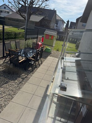 Une terrasse avec une table et des chaises, une maison de jeu pour enfants et, au loin, une vue sur d’autres maisons.