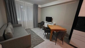 Un espacio habitable moderno y funcional con sofá, una pequeña mesa de comedor con sillas, televisor sobre un mueble bajo y una cocina con cubierta negra.