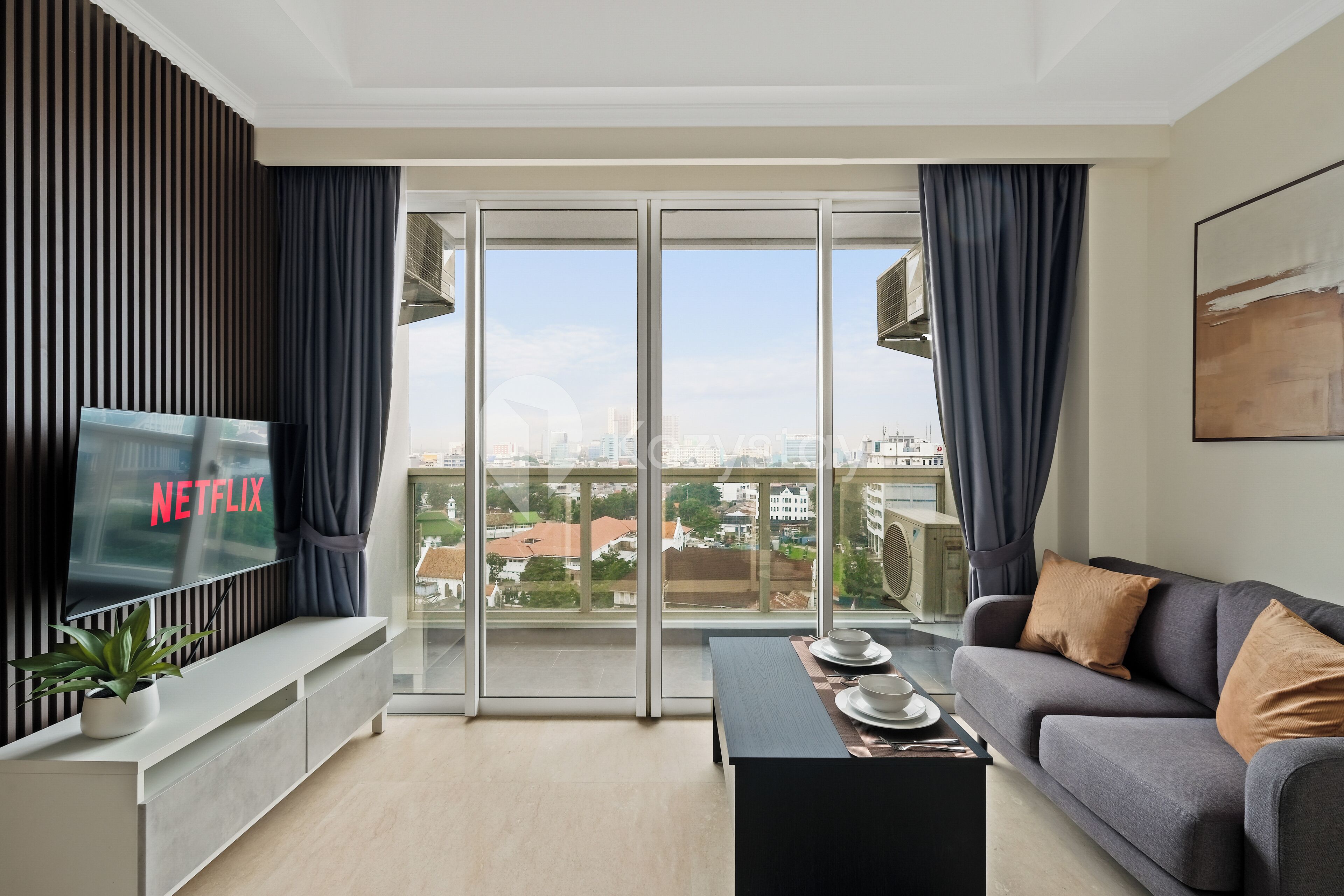 A modern living room with a flat-screen TV, a sofa, a coffee table, and a balcony with a view of the city.