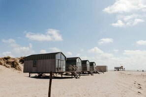 Des cabanes de plage aux façades vitrées et aux murs extérieurs en bois, disposées en rangée sur une plage de sable.