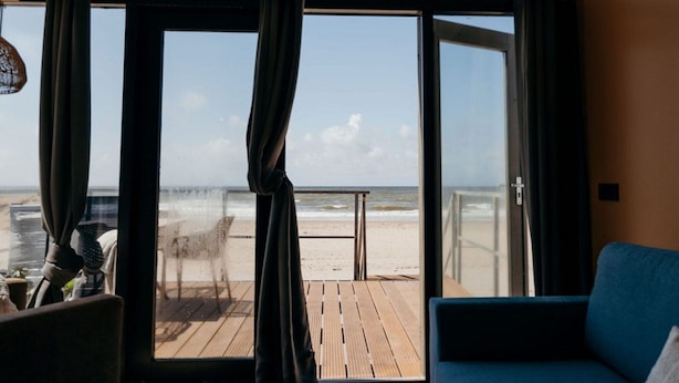 Een kamer met een groot raam dat uitkijkt over een strand. Buiten het raam ligt een houten terras, en in de kamer staat een blauwe bank.