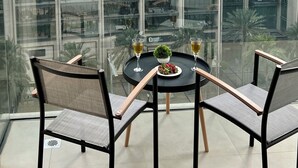 Un immeuble de grande hauteur doté d’un balcon en verre, aménagé avec deux chaises et une table, et agrémenté d’une petite plante en pot et de deux verres contenant une boisson.