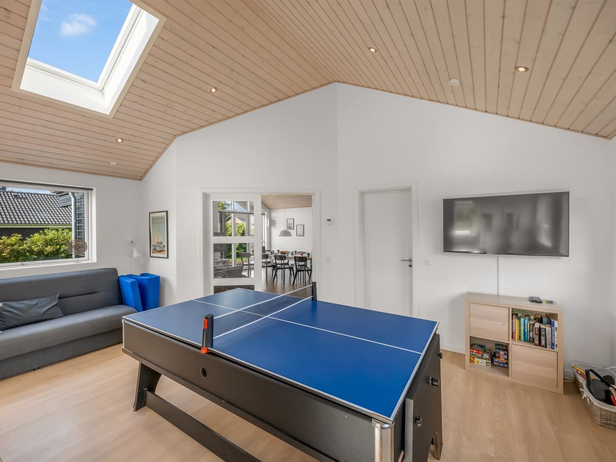 Un salón amplio con una mesa de ping pong, un sofá gris, una estantería y un televisor montado en la pared.