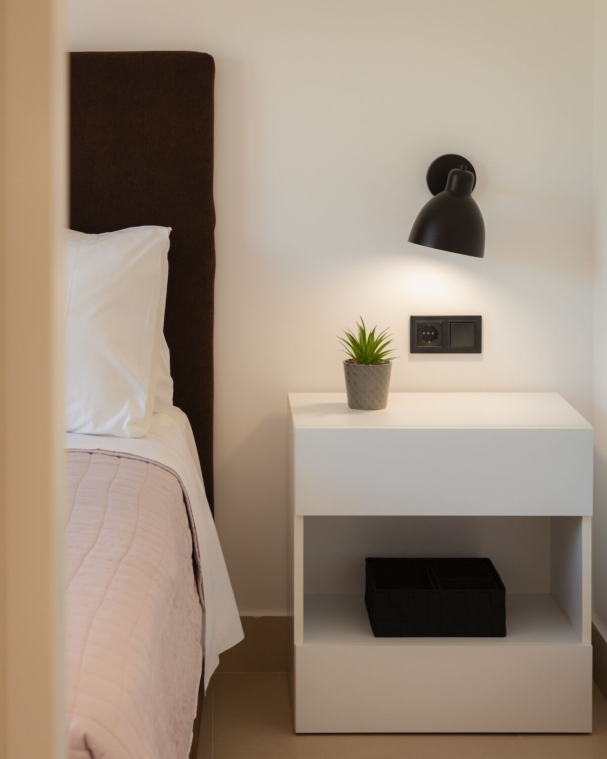 Ein Schlafzimmer mit einem Bett, einem weißen Nachttisch mit einer Pflanze, einer schwarzen Lampe und einer Steckdose.