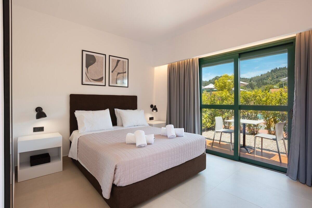 A modern bedroom with a large bed, two bedside tables, a nightstand, and a view of the outdoors through a sliding glass door.