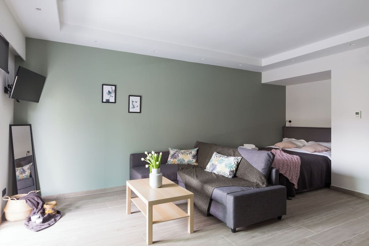 A modern, well-lit room with a grey sofa, a small wooden table, a bed with a grey headboard, and a TV mounted on the wall.