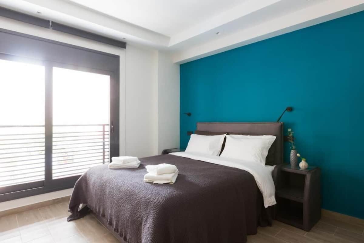 A modern hotel room with a large bed, a nightstand, a teal accent wall, and a view through a large window.