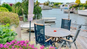 Eine Uferterrasse mit einem runden Tisch und Stühlen, umgeben von üppiger Bepflanzung und einem Kanal mit Booten.