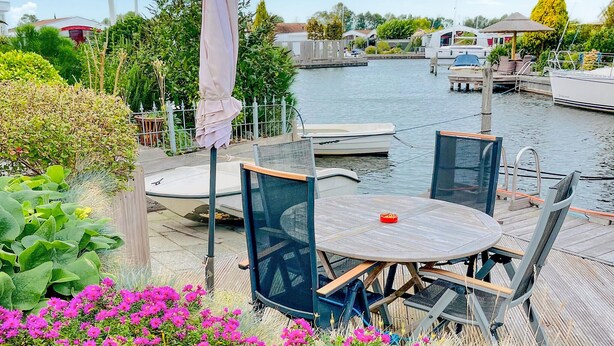 Eine Uferterrasse mit einem runden Tisch und Stühlen, umgeben von üppiger Bepflanzung und einem Kanal mit Booten.