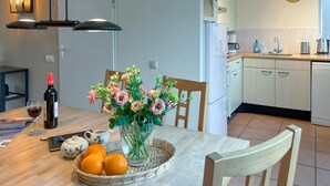 Eine moderne Küche mit weißen Schränken, einer Kaffeemaschine und einem Spülbecken. Ein Esstisch ist mit einer Blumenvase, einer Weinflasche und einem Korb mit Orangen dekoriert.