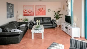 Un salon avec un canapé en cuir noir, une table basse blanche ornée de fleurs, un canapé gris, un fauteuil à rayures, un téléviseur posé sur un meuble blanc et des œuvres d’art accrochées aux murs.