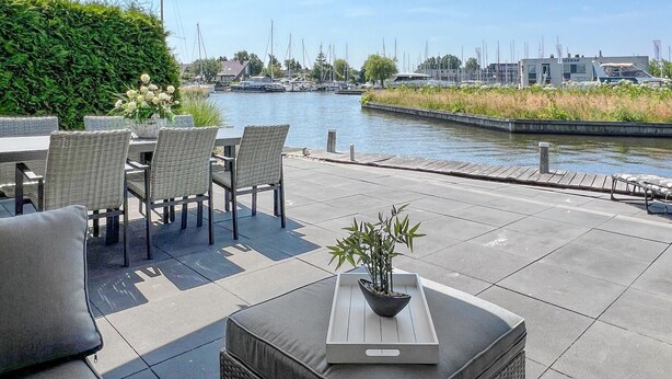 Une terrasse extérieure donnant sur un canal, avec une table et des chaises, un pot de fleurs et un petit bateau amarré à proximité.