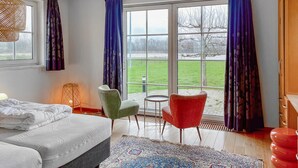 A bedroom with a bed, a green chair, a red chair, a small table, and a view of a green field through large windows.