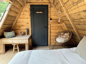 A cozy attic bedroom with a dark door, a small wooden table, a wicker chair with cushions, and a bed with a white comforter. - Tipi d'HamiH" unusual accommodation (Uzès)