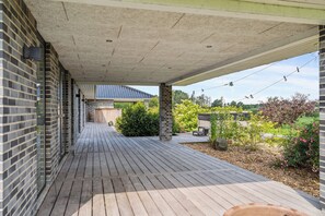 Une terrasse en bois couverte, avec un mur en briques, un toit recouvert de tuiles bleues en arriĂšre-plan, et un espace jardin agrĂ©mentĂ© de plantes et dâun banc.