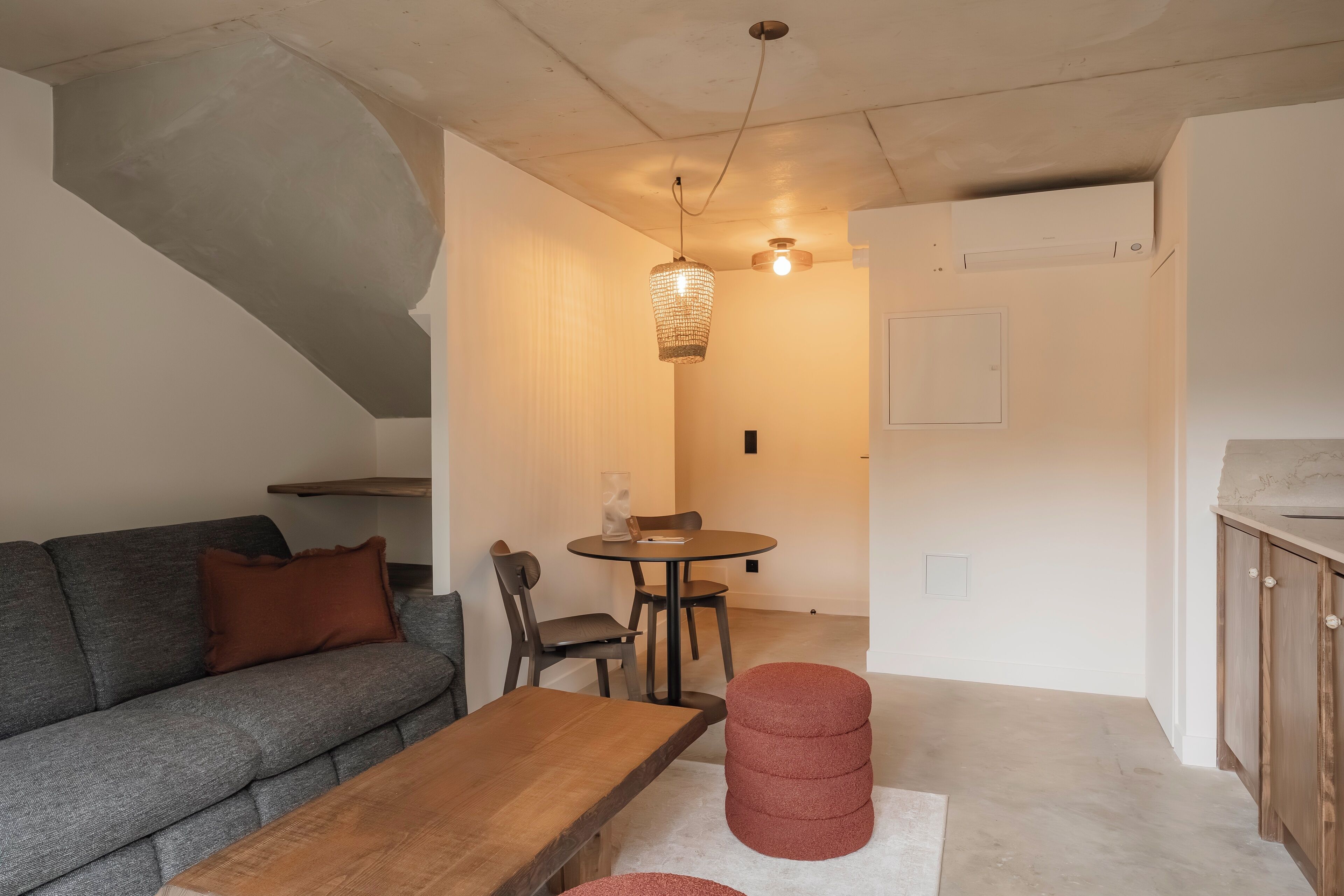 A living room with a sofa, a wooden table, and a red ottoman. There is a dining area with a table and chairs, and a kitchen area with cabinets.