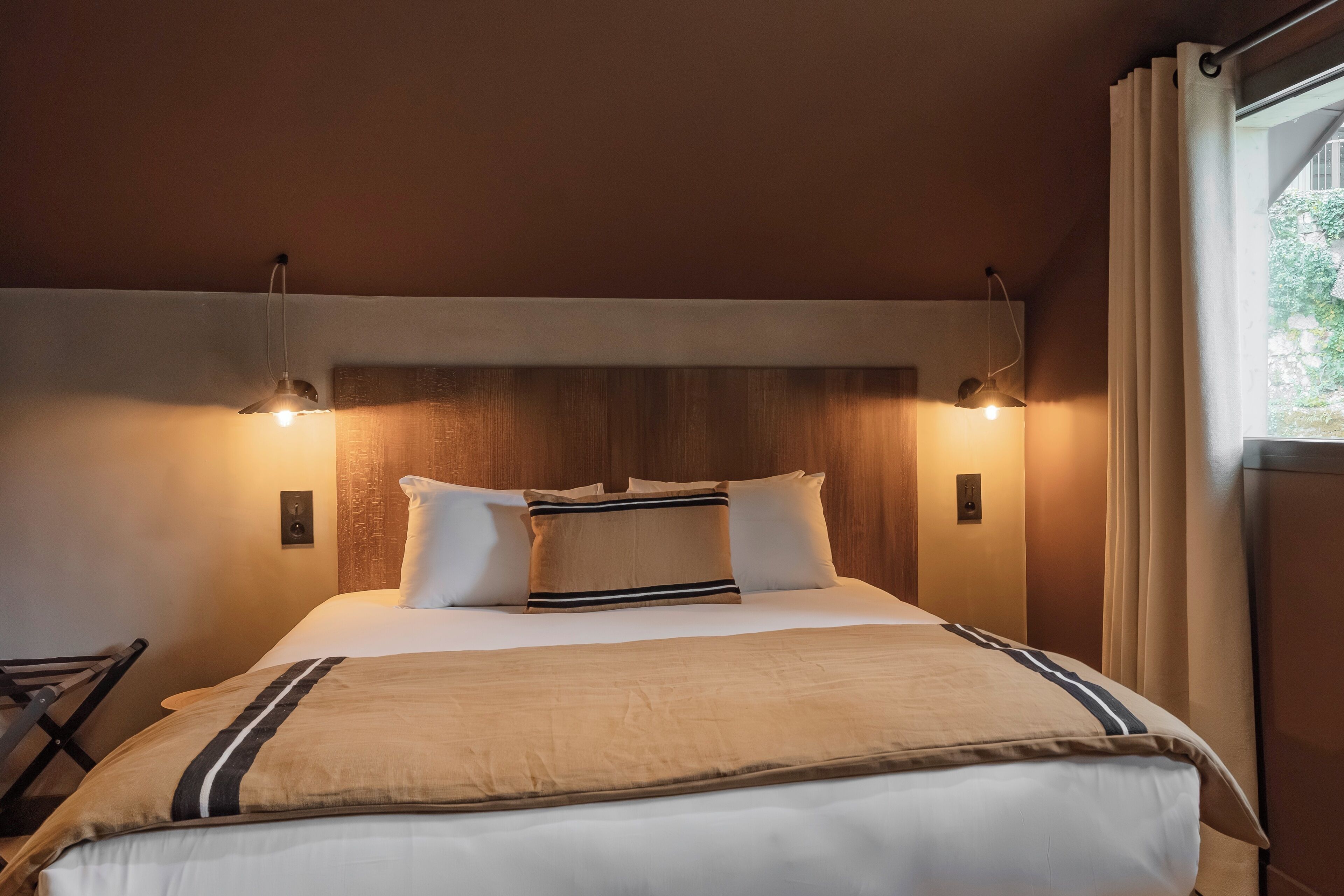 Un lit bien fait, recouvert d’une couvre-lit à rayures, avec deux lampes de chevet et une vue sur un jardin par une fenêtre ornée de rideaux.