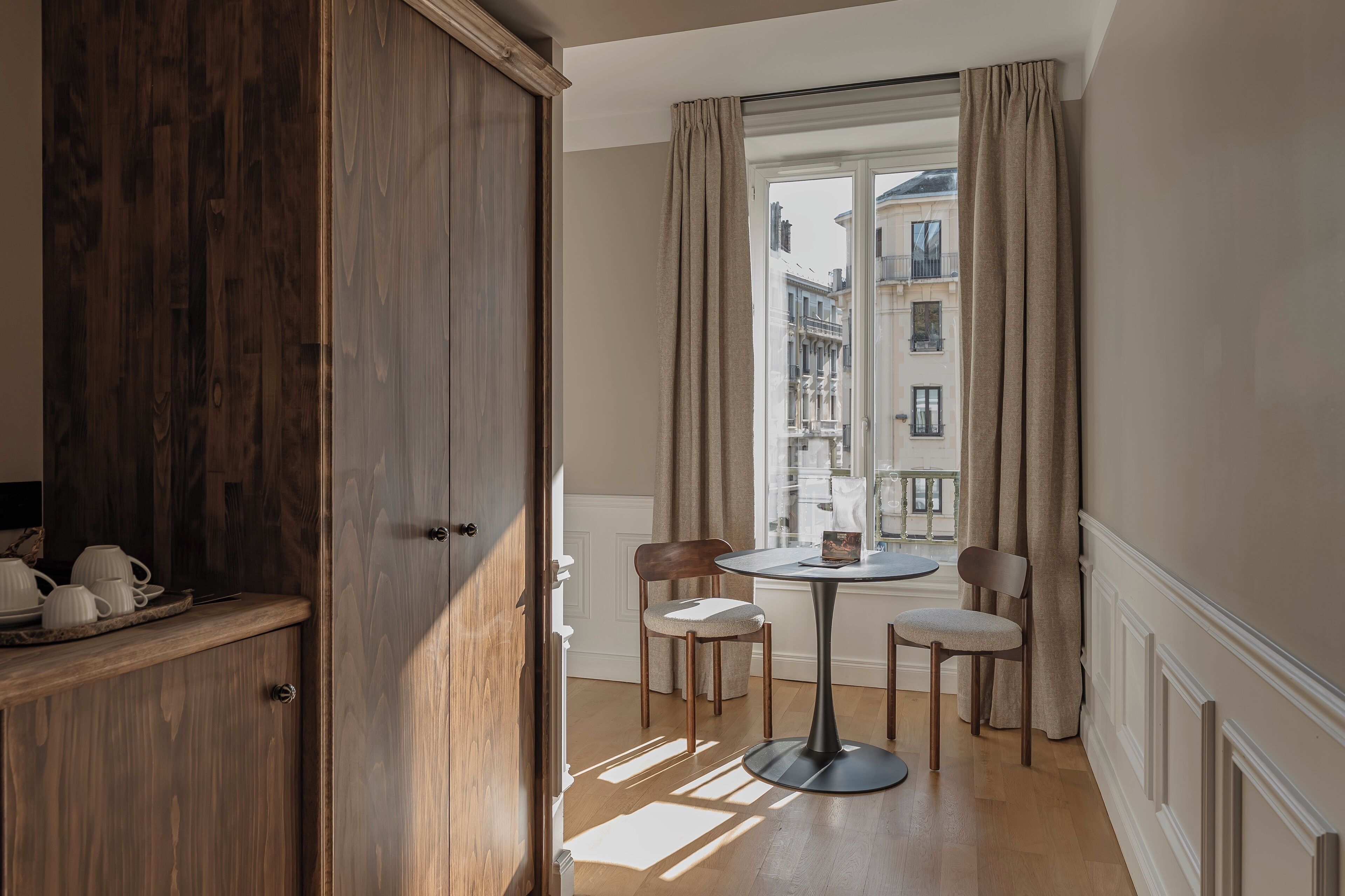 Une pièce avec une armoire en bois, une table ronde entourée de chaises et une fenêtre avec des rideaux.