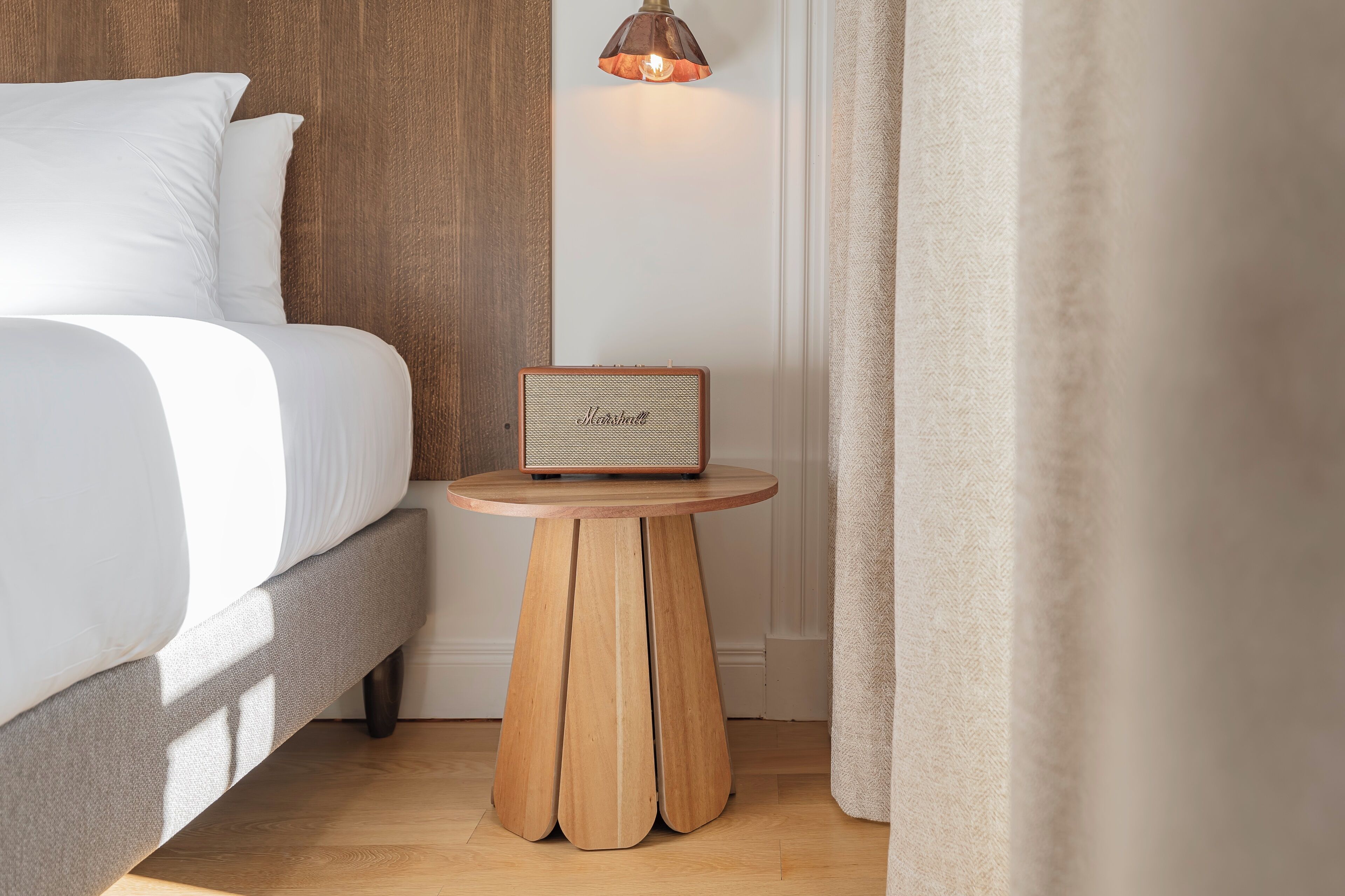 A neatly made bed with white linens, a wooden side table with a vintage-style radio, and a hanging light fixture.