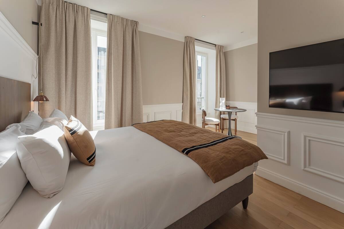 A hotel room with a large bed, a TV mounted on the wall, and a small table with chairs.