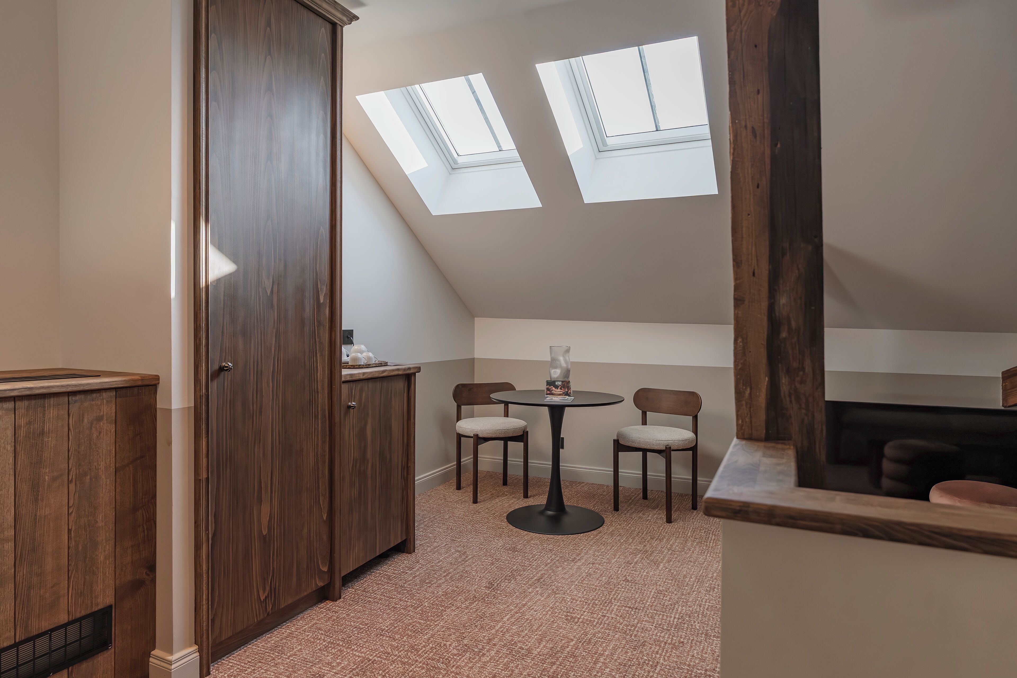 Una habitación con un armario de madera, una mesa redonda con sillas, un tragaluz y piso alfombrado.