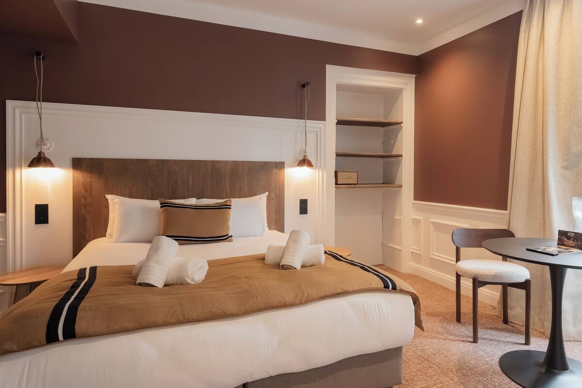 A neatly made bed with a brown headboard, two bedside tables, a small round table with a chair, and a built-in shelving unit.