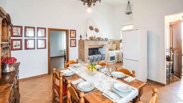 A dining area with a wooden table set for six, surrounded by chairs. A kitchen with a stove and refrigerator is visible in the background.