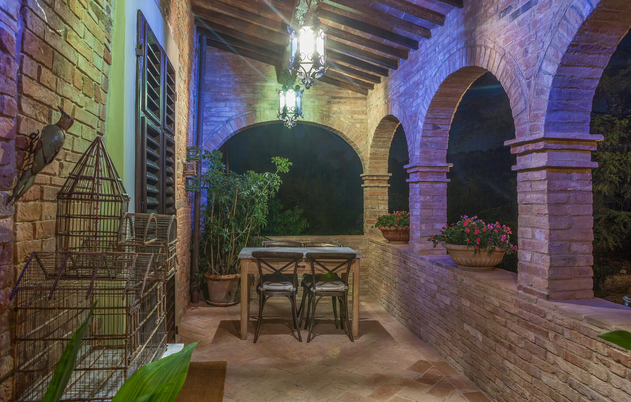 Un patio al aire libre con una mesa y sillas, rodeado de muros y arcos de ladrillo, y decorado con macetas y jaulas de pájaros.