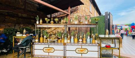 A kiosk with a tiled counter and various decorative items, set against a stone wall and under a metal roof.