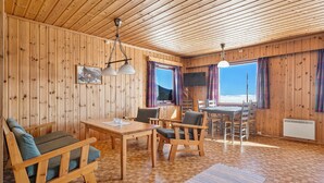 Interior de una cabaña de madera con una mesa de comedor, sillas, un sofá, una televisión y vistas al exterior.