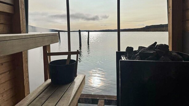 Un banco de madera y un cubo junto a una ventana con vistas a una masa de agua tranquila y una costa lejana.