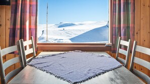 Una habitación con una mesa de madera y dos sillas, una ventana con cortinas a cuadros y vistas a un paisaje nevado.