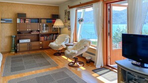Un salon chaleureux avec une grande fenêtre, une bibliothèque, un fauteuil confortable et un téléviseur.