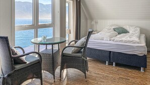 A room with a bed, two chairs, a small table, and a view of the sea through large windows.
