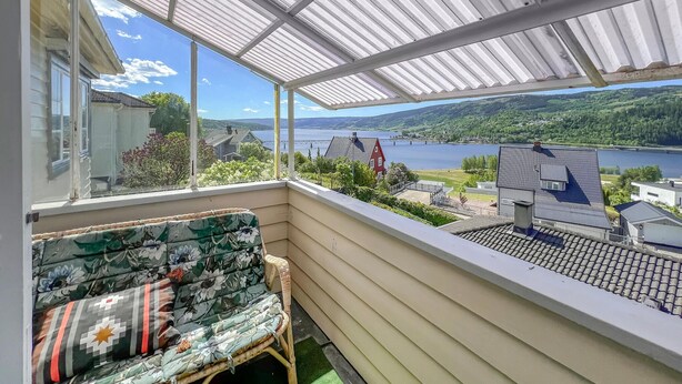 A balcony with a view of a lake and a bridge, a wicker chair, and a window with a view of a house.
