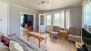 A living room with a sofa, two armchairs, a coffee table, and a TV. There is a door leading to another room and a window with curtains.