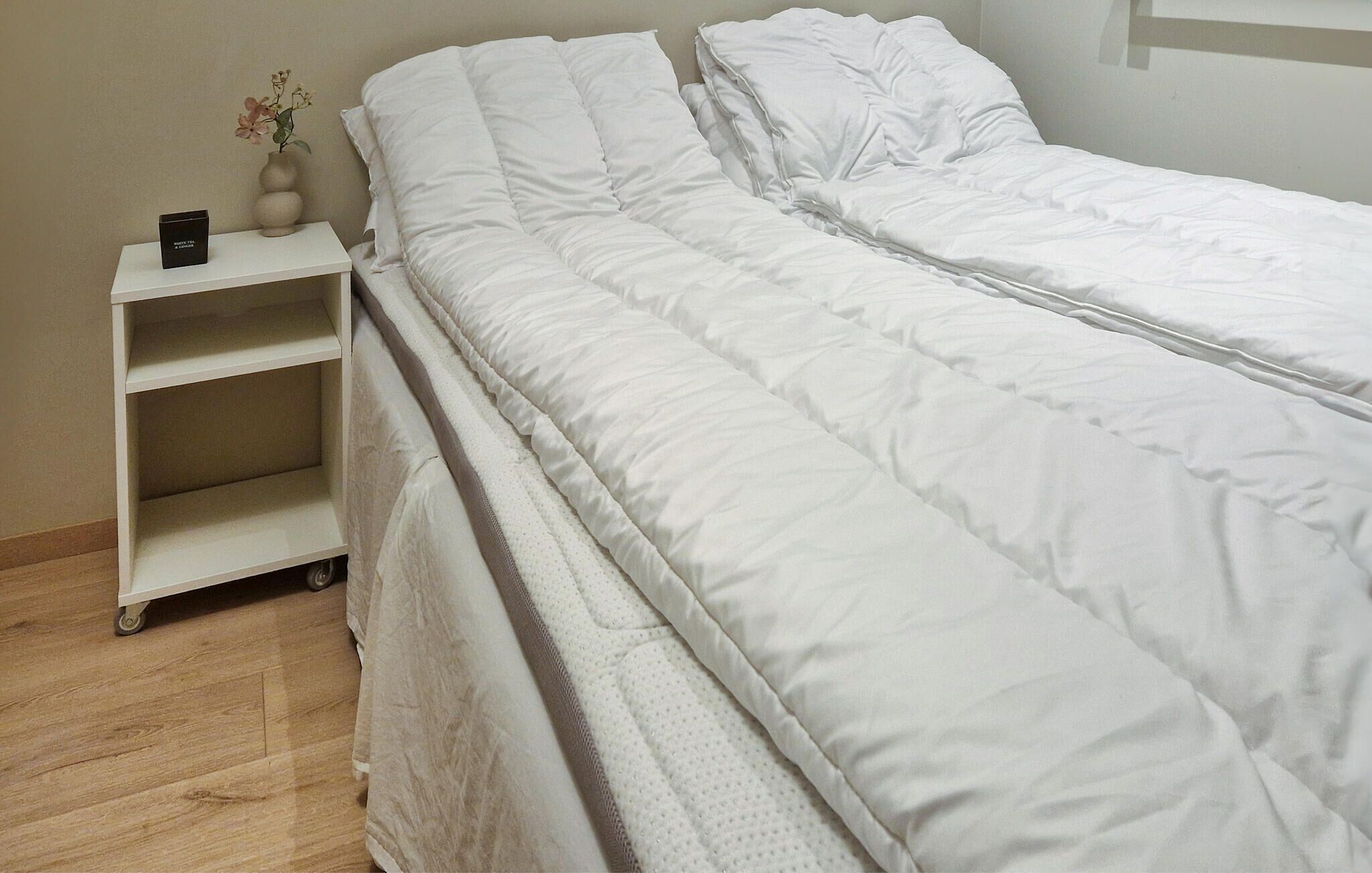 A neatly made bed with white bedding, a small bedside table with a vase of flowers, and a shelf unit with a box on it.