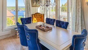 Un espace repas avec une table blanche, six chaises bleues à revêtement en tissu, un lustre et une vue sur une étendue d’eau grâce à de grandes fenêtres.
