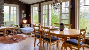 A cozy dining area with a wooden table and chairs, a stone fireplace, and large windows offering a view of trees.