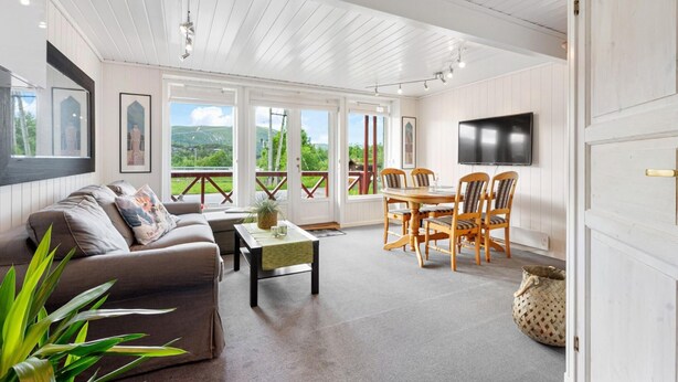 Ein geräumiges Wohnzimmer mit Sofa, Couchtisch und Essbereich mit Stühlen. Ein Fernseher ist an der Wand befestigt und ein großes Fenster bietet einen Blick ins Freie.
