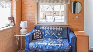 Una habitación acogedora con detalles de madera, una ventana que da a una terraza de madera, una mesita con una lámpara y un sofá con cubierta acolchada.