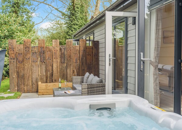 A hot tub with a wooden deck, wicker furniture, and a glass door leading to a bedroom.