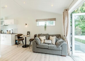 Ein modernes Wohnzimmer mit grauem Sofa, einem kleinen Tisch, einer Küchenzeile und einem großen Fenster mit Vorhängen.