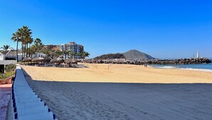 A beach with a staircase leading down to the sand, palm trees, a building with a thatched roof, and a lighthouse in the distance.