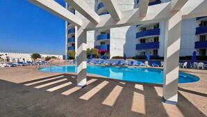 A pool area with multiple lounge chairs and umbrellas, a clear blue sky, and a building with balconies in the background.
