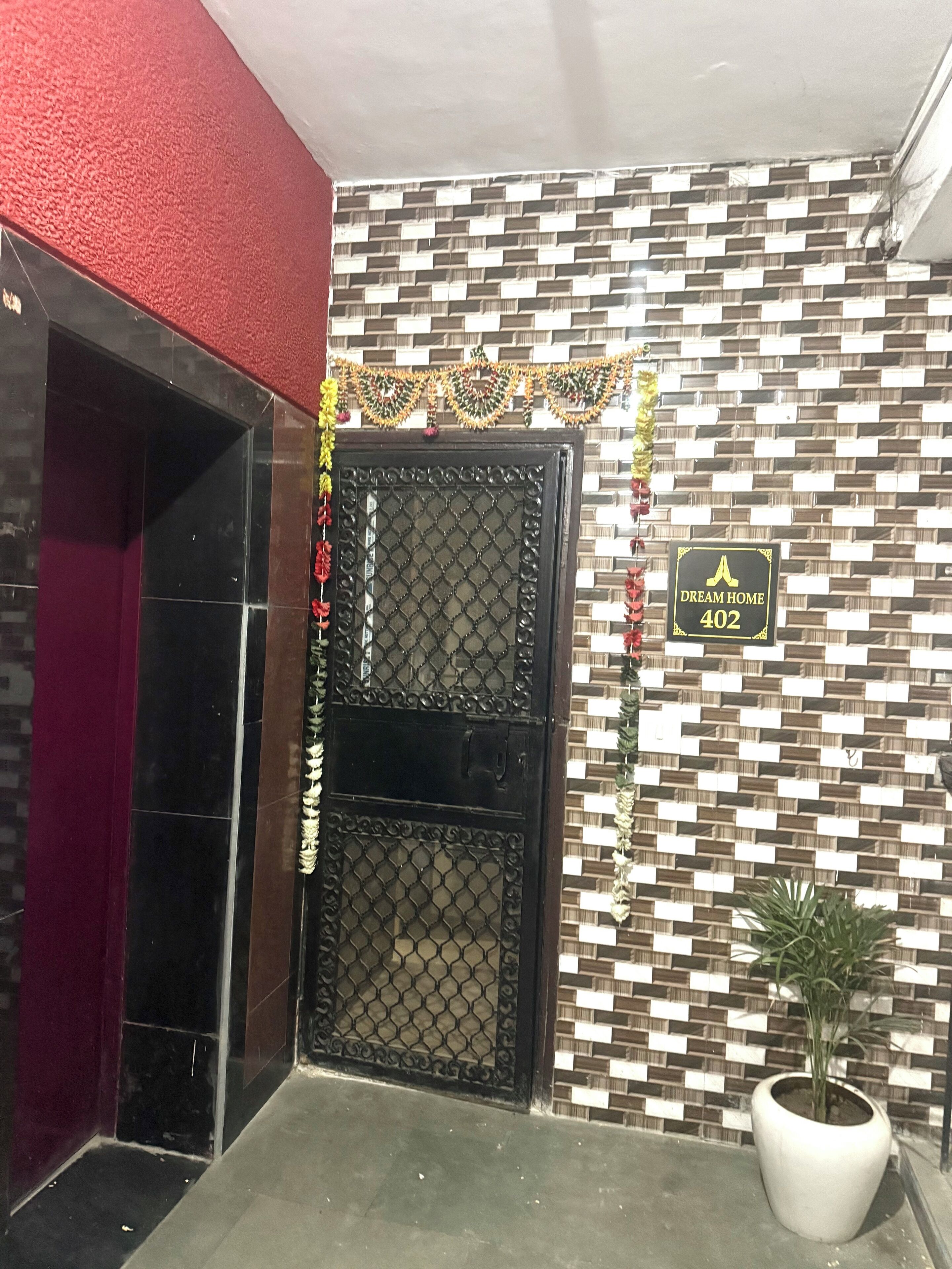 A black double door with a decorative sign above it, a potted plant to the right, and a patterned wall in the background.