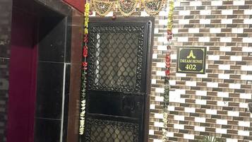 A black double door with a decorative sign above it, a potted plant to the right, and a patterned wall in the background.